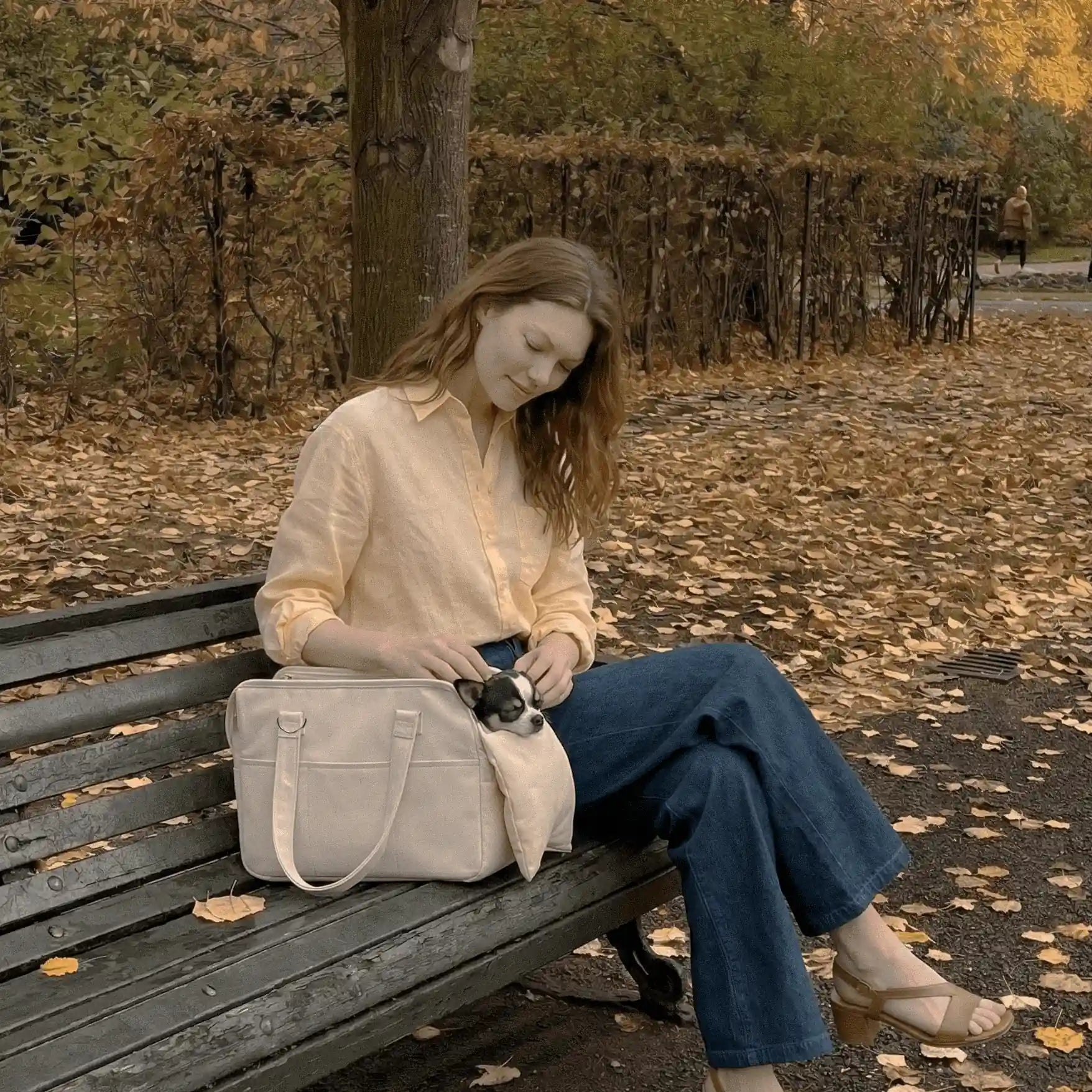 A model is sitting on a park bench, petting a sleeping Chihuahua inside a pet carrier bag